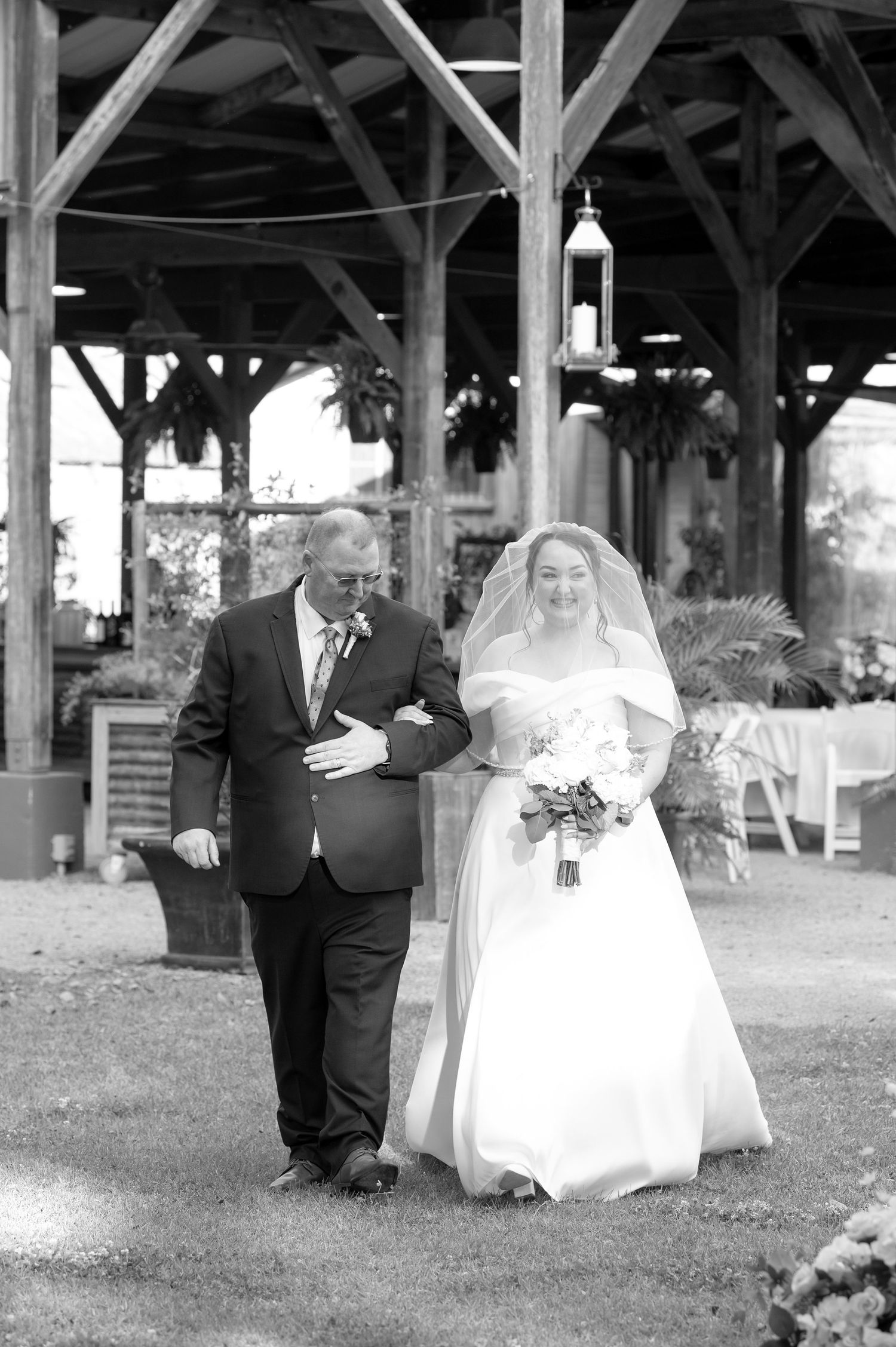 Black and white photo of a bride in white gown and veil walking down the aisle with her father at an outdoor wedding.