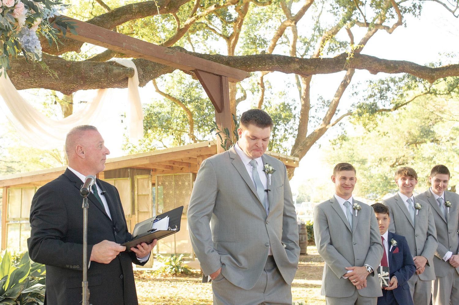 Groom in gray suit stands at outdoor wedding ceremony with groomsmen and officiant under oak tree.