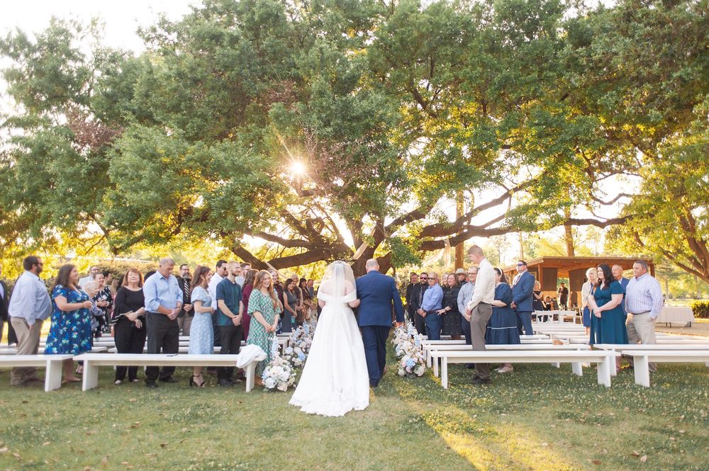 Bride and groom stand before guests at an elegant outdoor wedding ceremony under a large oak tree at golden hour.