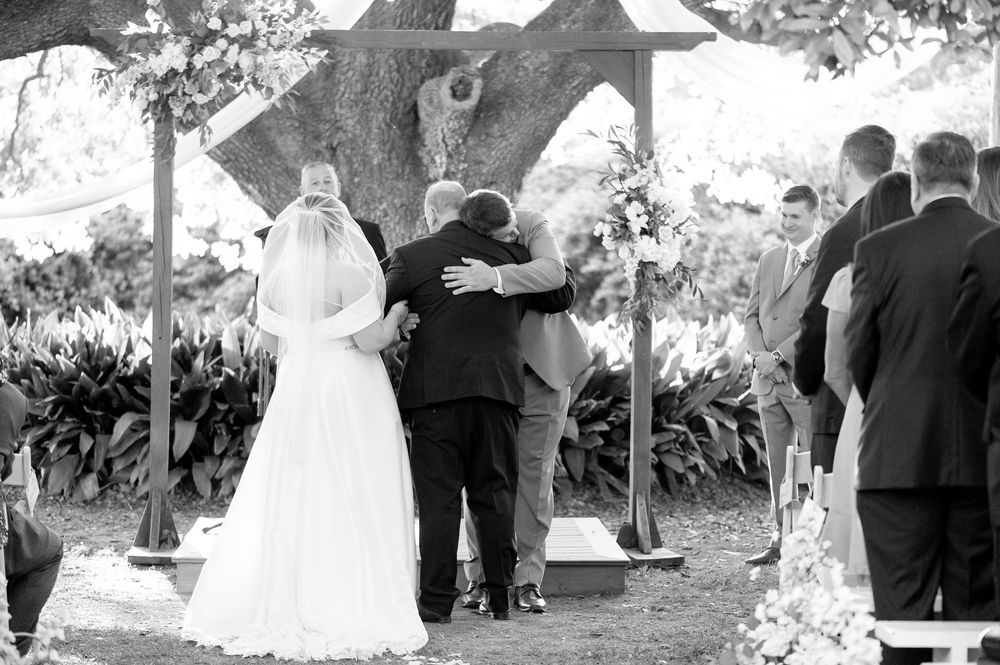 Black and white outdoor wedding ceremony with bride, groom, and officiant embracing under a wooden arch.