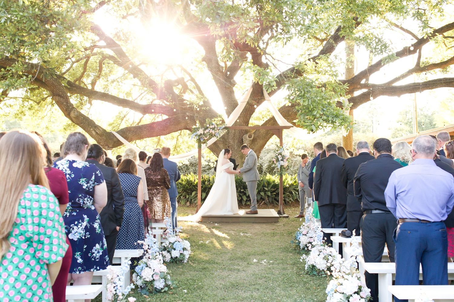 Outdoor wedding ceremony under large oak tree with white chairs and floral arrangements lining the aisle.