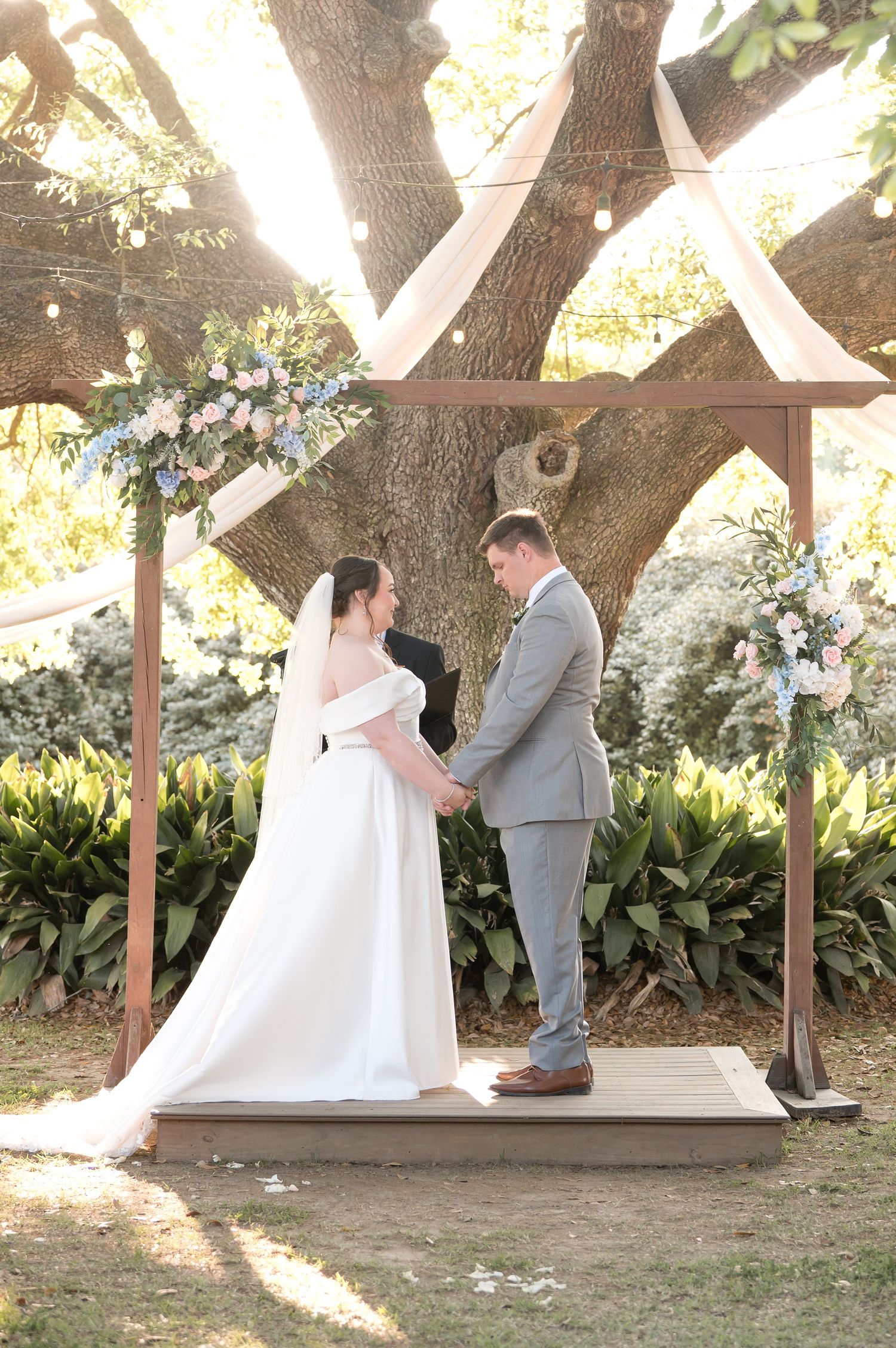 Bride and groom hold hands at outdoor wedding altar under large tree with floral arrangements and wooden pergola.