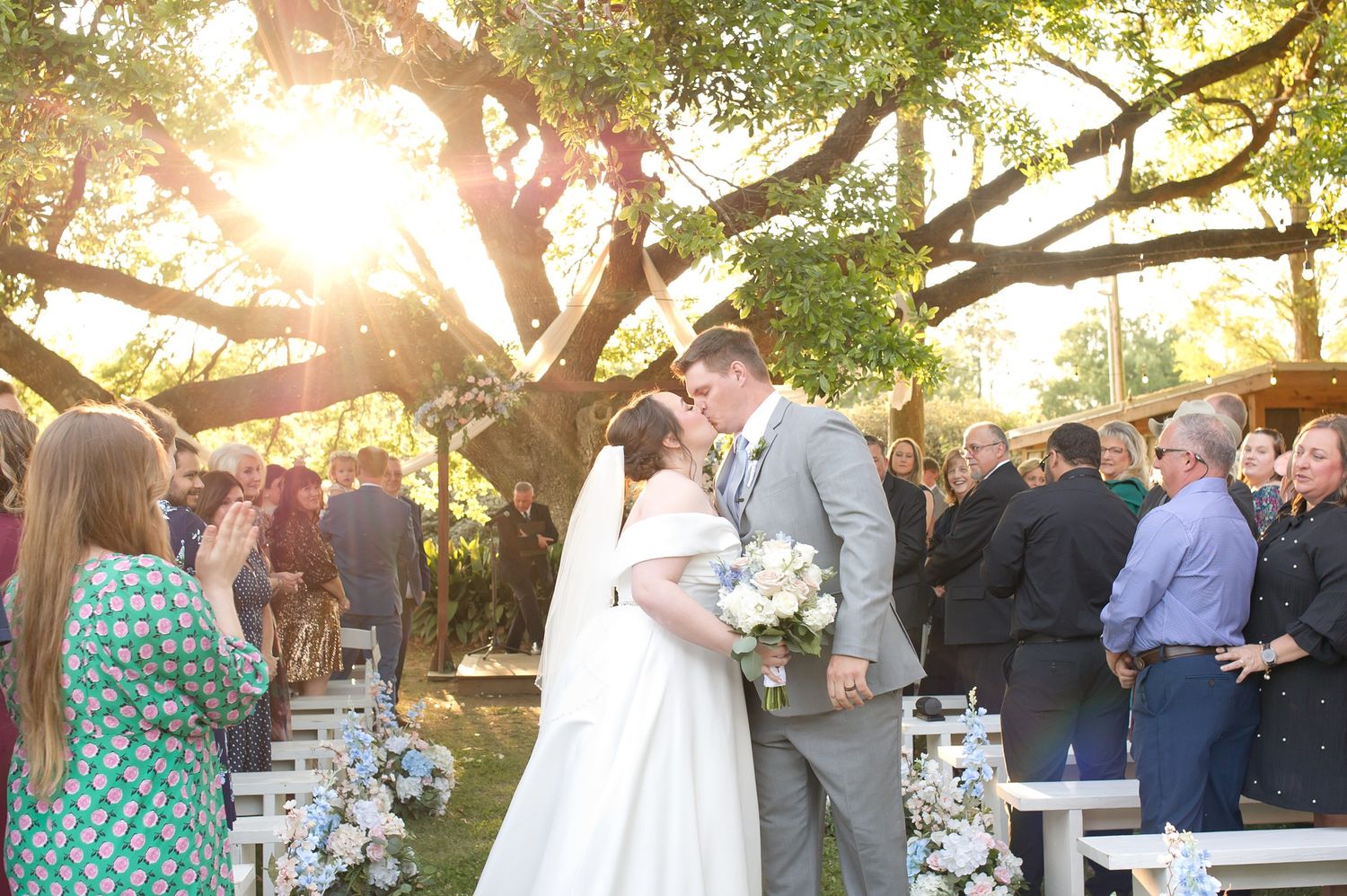 Bride and groom share first kiss at outdoor wedding ceremony under sunlit oak tree with guests applauding.