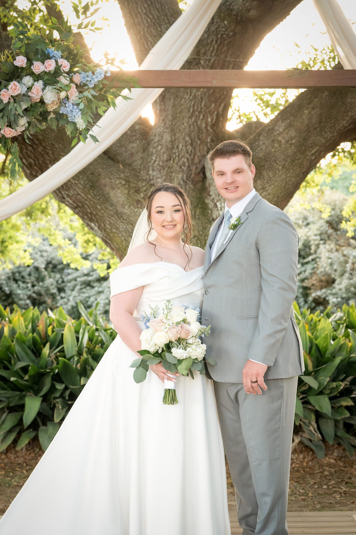 Bride and groom pose under floral arch with white draping and string lights at outdoor wedding ceremony.