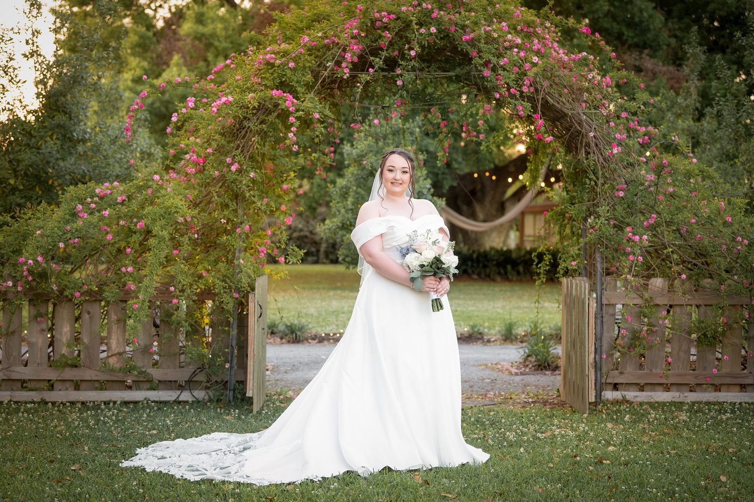 Bride in white gown holding bouquet stands under floral arch with pink flowers at outdoor wedding venue.