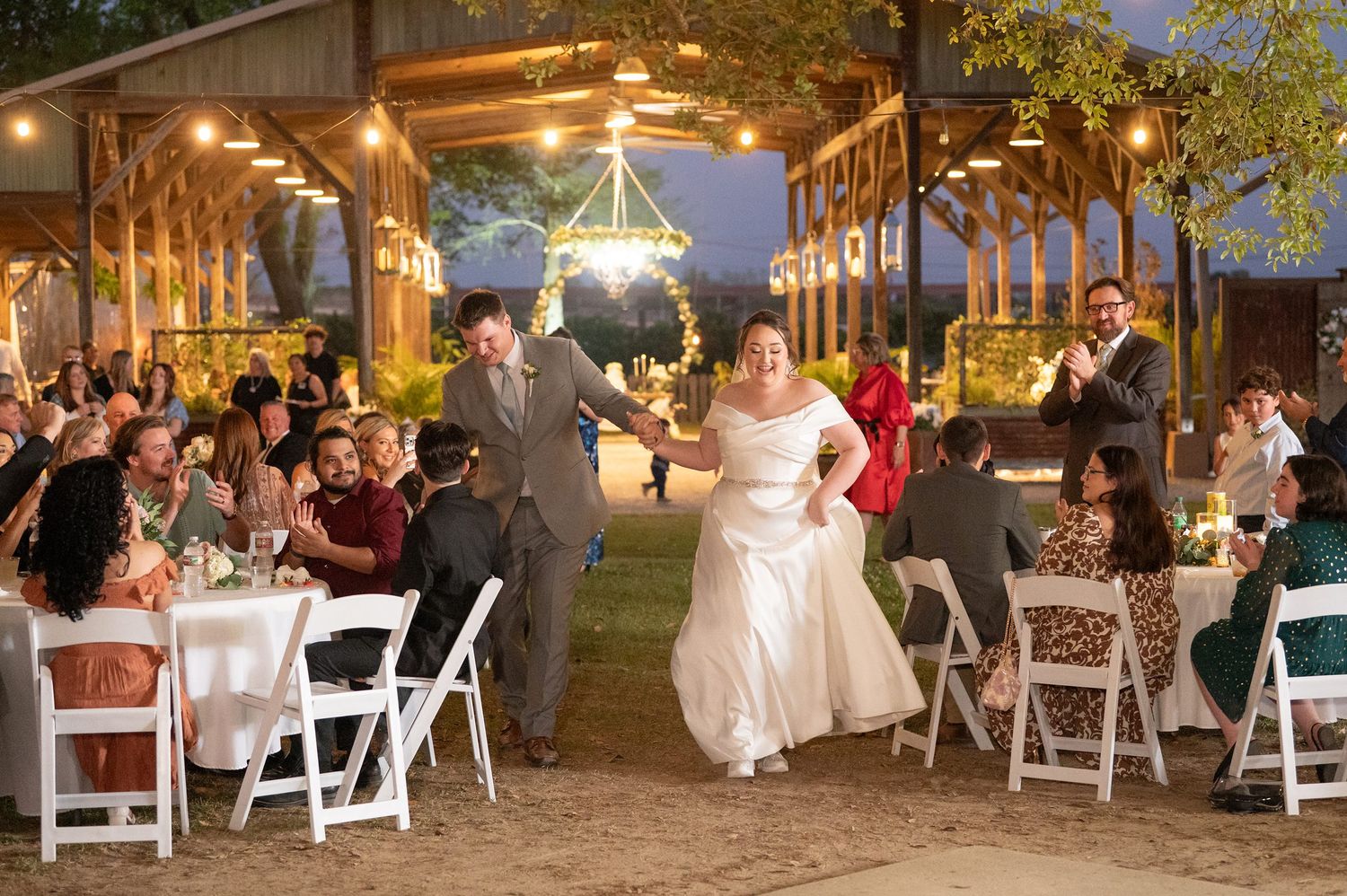 Bride and groom make grand entrance at outdoor evening wedding reception under warm string lights.
