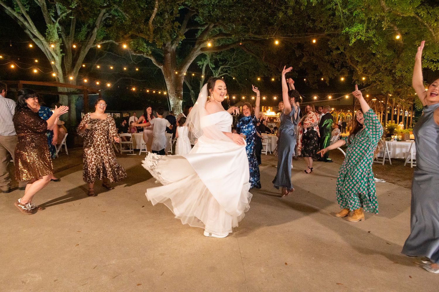 Bride twirling in white gown on outdoor dance floor at night wedding reception with string lights.