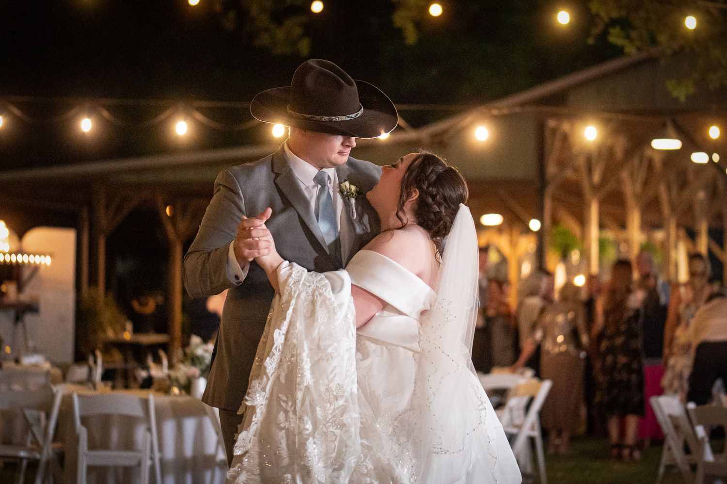 Bride and groom share first dance outdoors at night under string lights at rustic country wedding reception.