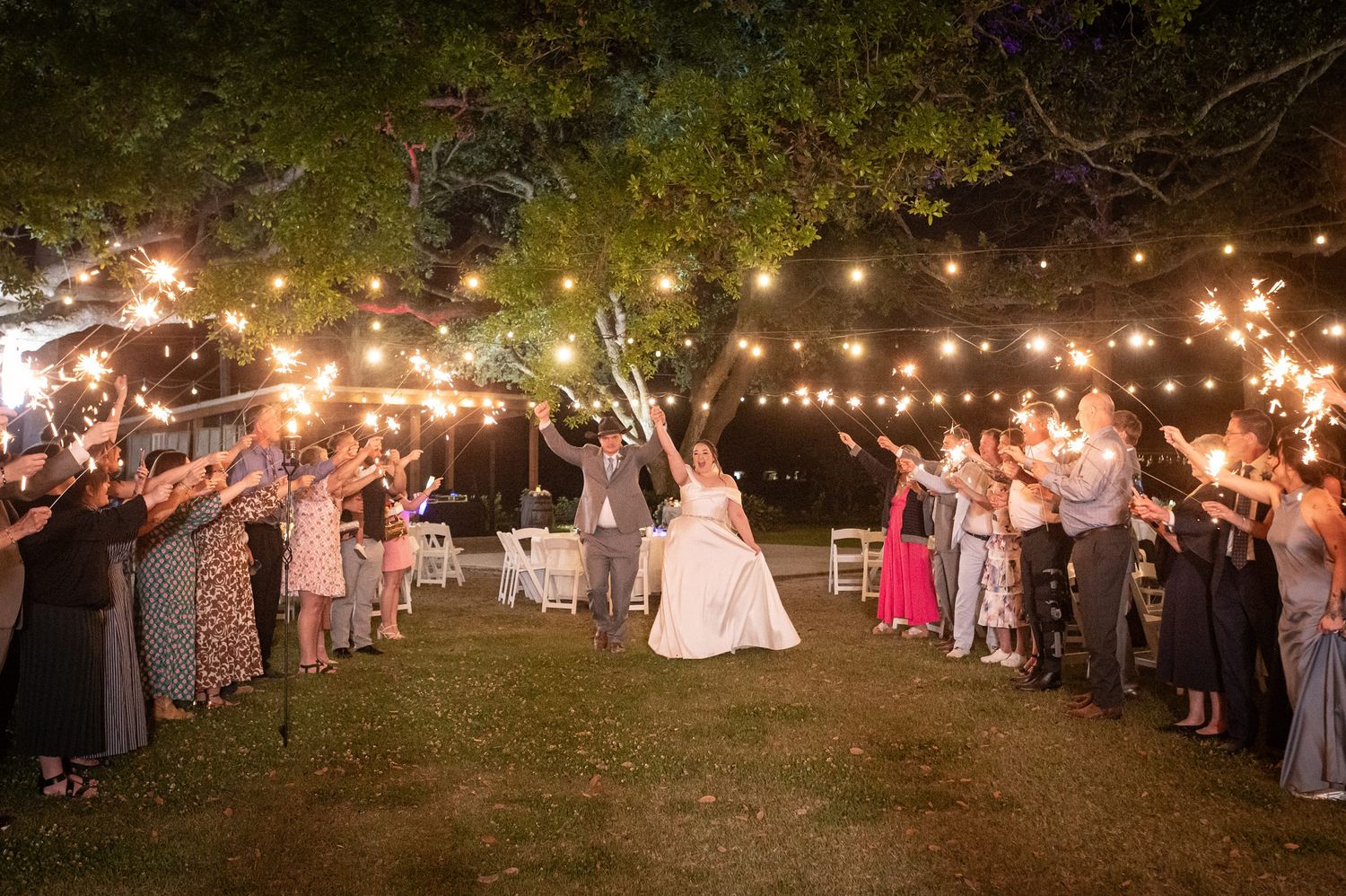 Bride and groom exit through sparkler tunnel surrounded by cheering guests at nighttime wedding celebration.
