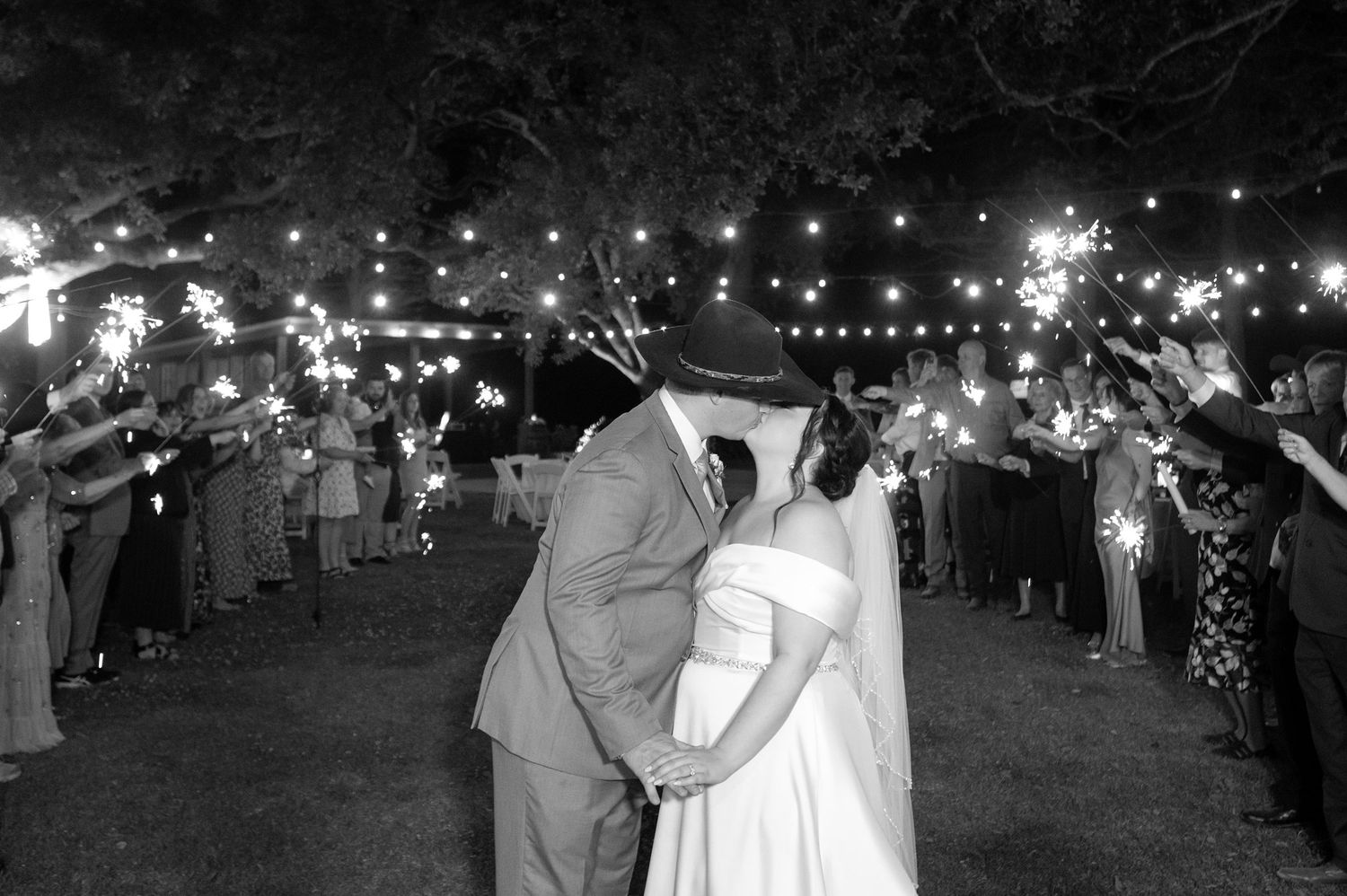 Black and white photo of newlyweds kissing during sparkler exit at outdoor wedding reception at night.