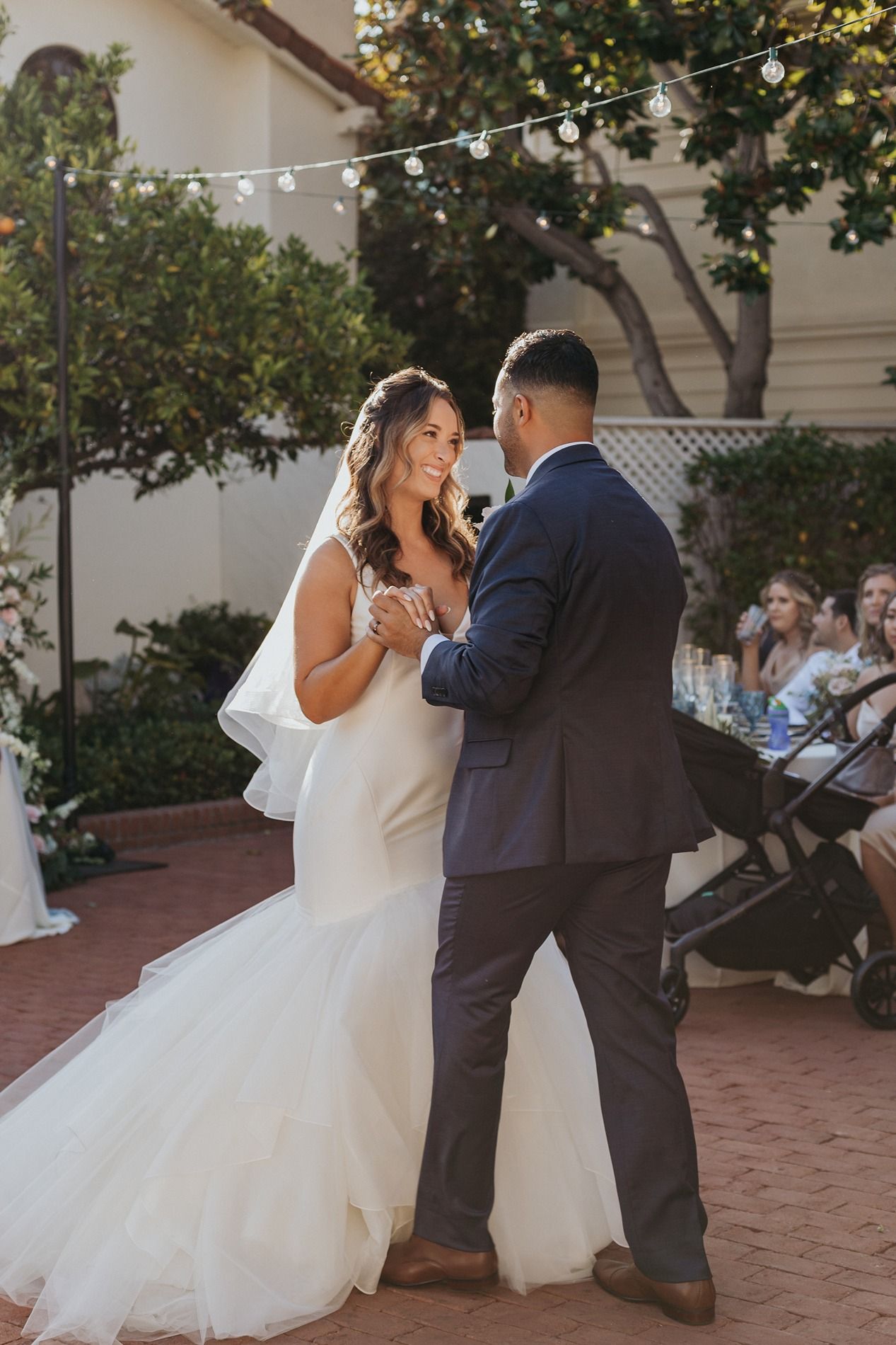 Bride and groom share their first dance outdoors at a wedding reception, with string lights and guests in the background.