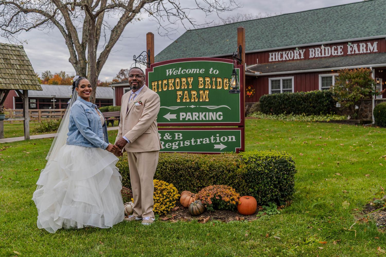 Couple in wedding attire posing by Hickory Bridge Farm welcome sign with red barn in background.