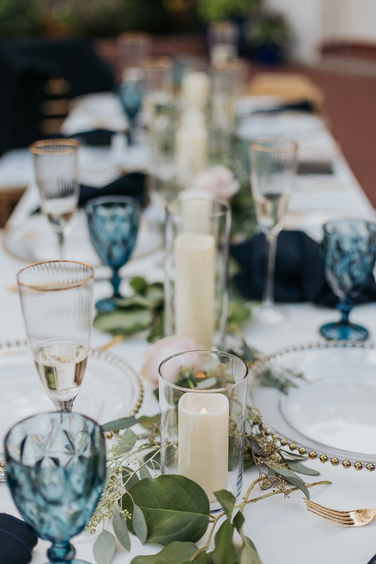 Elegant wedding table setting with blue goblets, gold-rimmed glasses, white candles, and eucalyptus greenery on white linen.