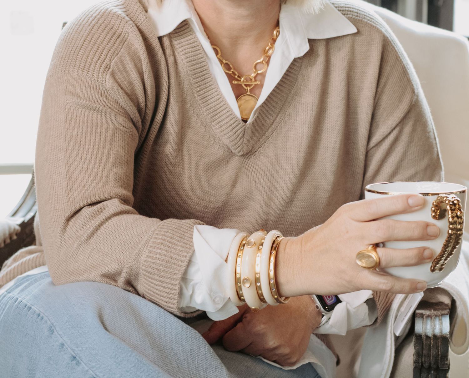 Woman wearing gold chain necklace, layered bracelets, and a beige V-neck sweater over white shirt, holding a white mug.