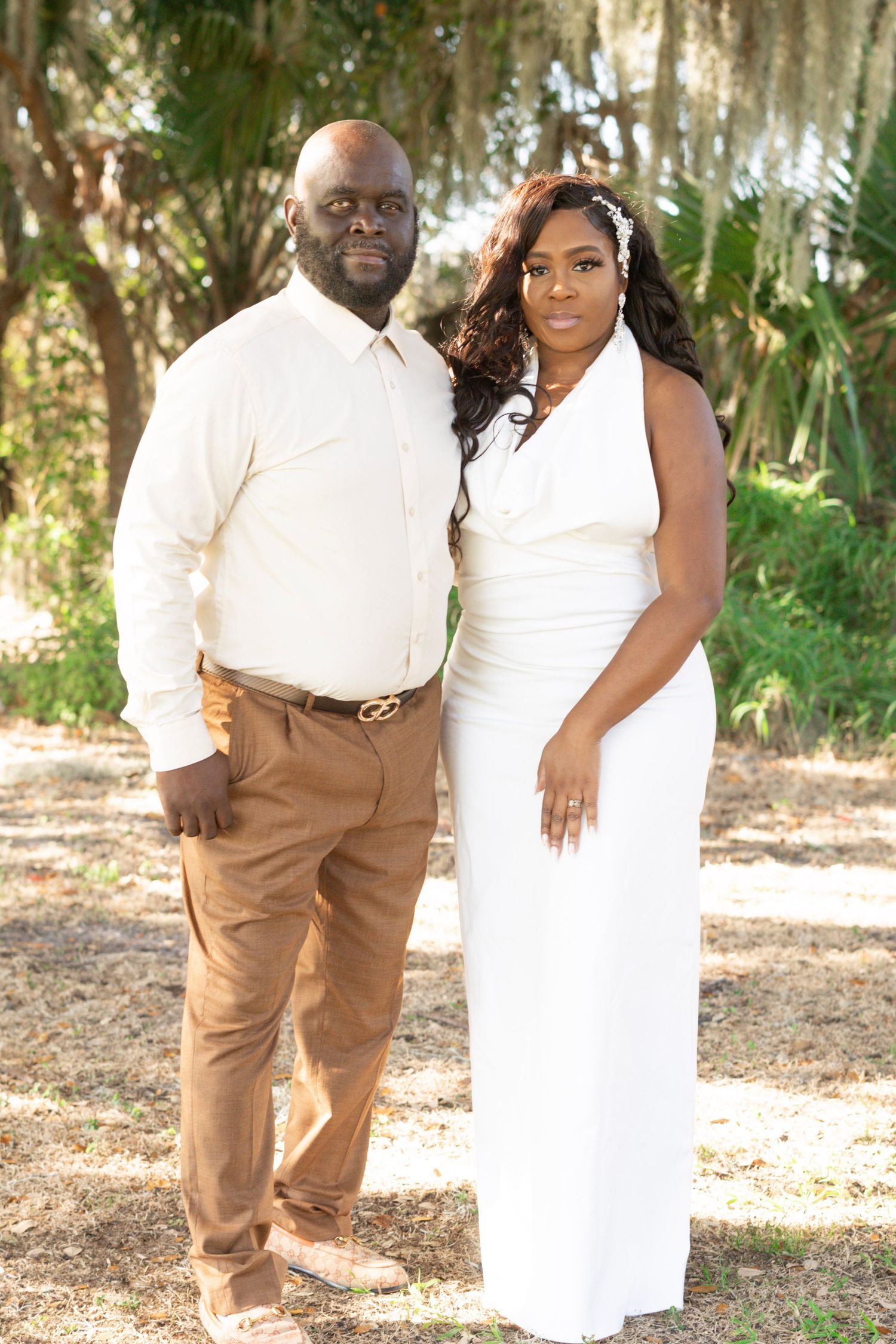 A couple poses together outdoors in coordinated white and tan outfits against a natural wooded backdrop.
