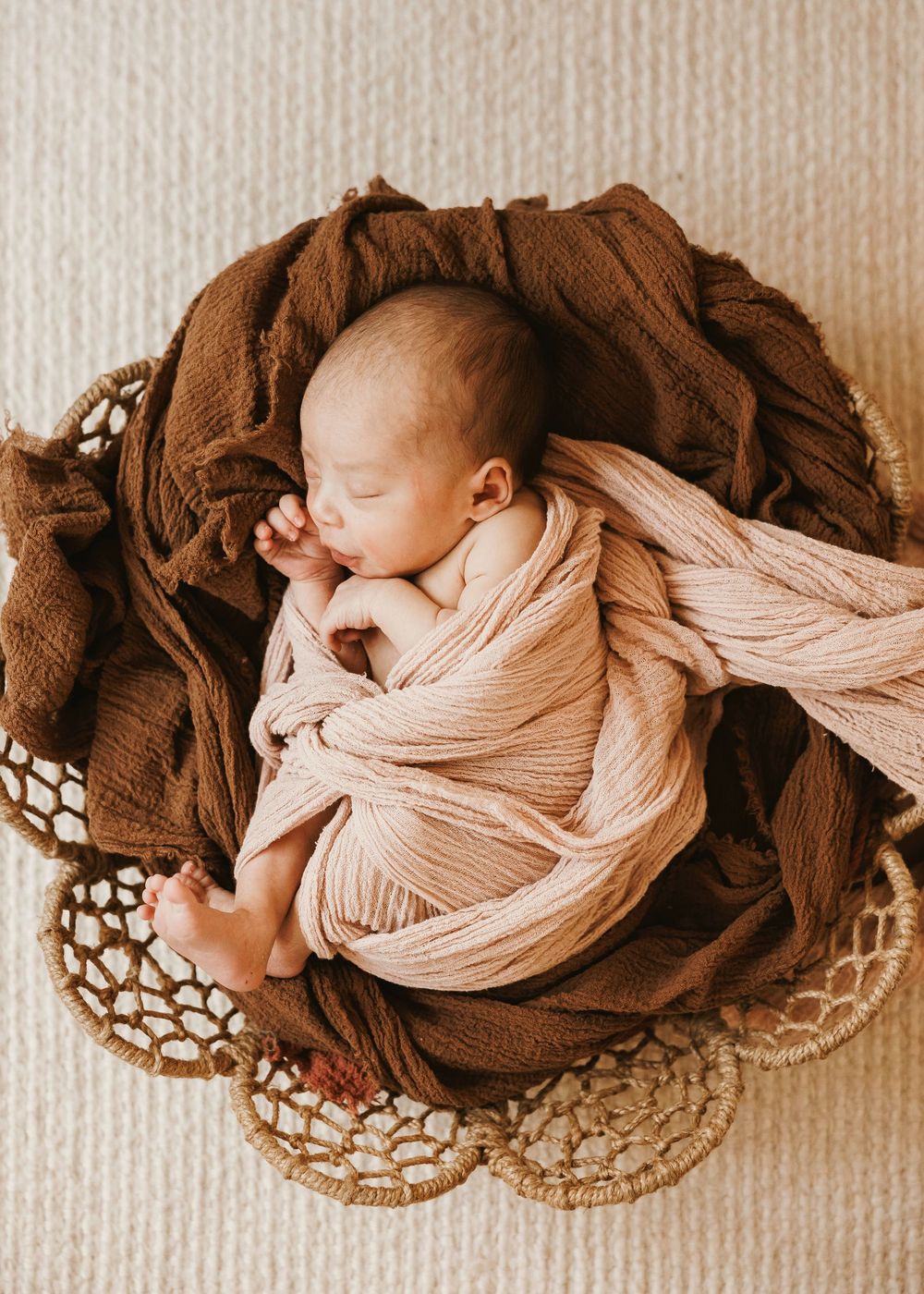 Sleeping newborn baby swaddled in pink and brown wraps resting in a woven basket.