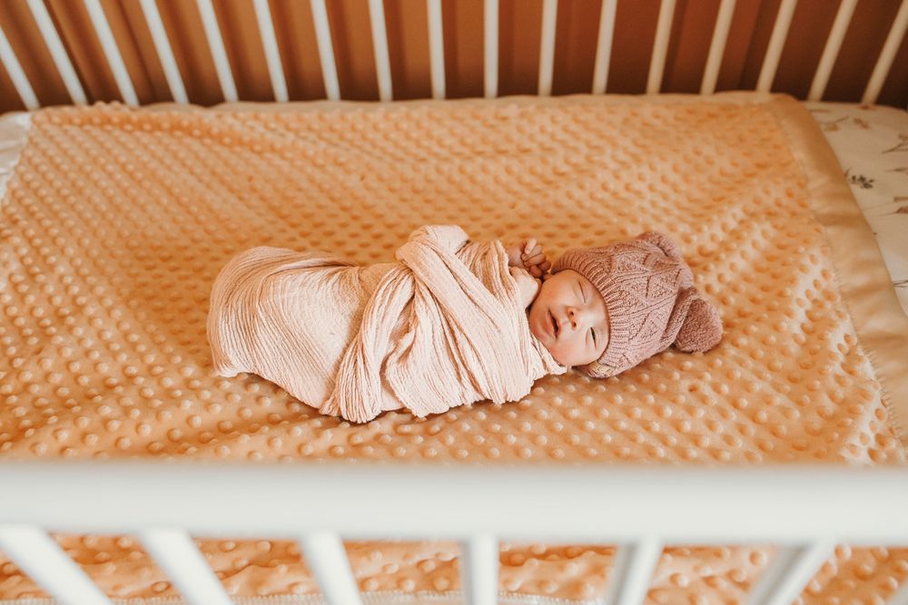 Newborn baby swaddled in a white wrap and pink bonnet lying peacefully in a white crib on a peach blanket.