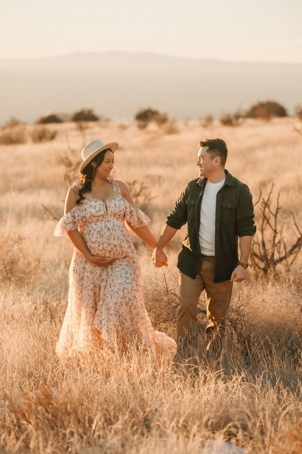 A pregnant couple holds hands while walking through a golden field at sunset.