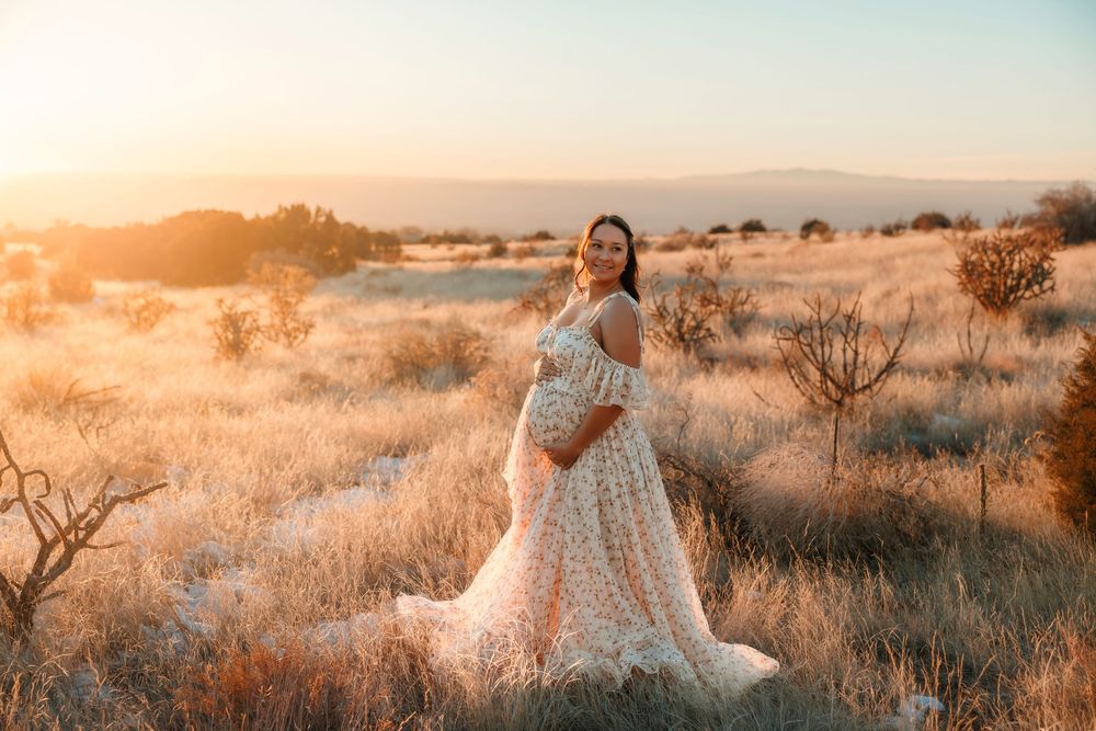 Pregnant woman in a flowing floral gown standing in a golden field at sunset with mountains in the background.