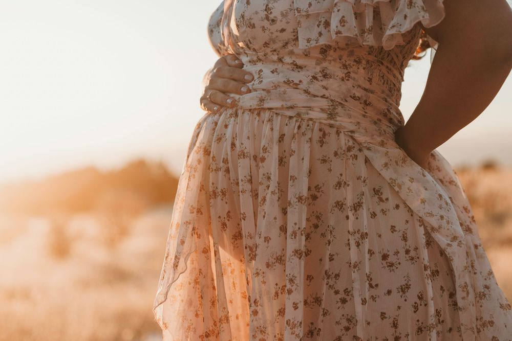 Close-up of a pregnant woman in a floral dress holding her baby bump during a golden hour outdoor photoshoot.