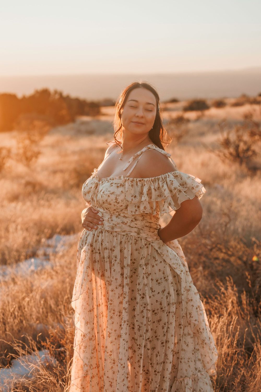 Pregnant woman in an off-shoulder floral dress posing at sunset in a dry golden field with mountains afar.