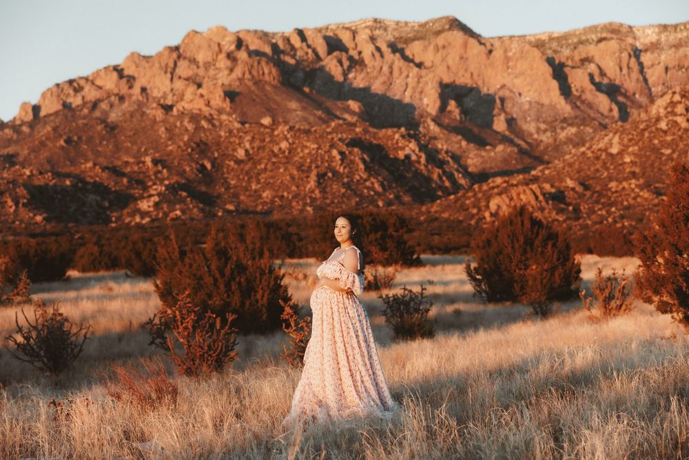 Pregnant woman in a floral maternity gown standing in a desert field with dramatic red mountains at sunset.