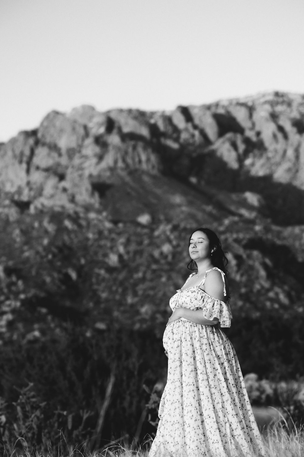 Black and white portrait of a pregnant woman in a floral dress cradling her bump with rocky mountains behind her.