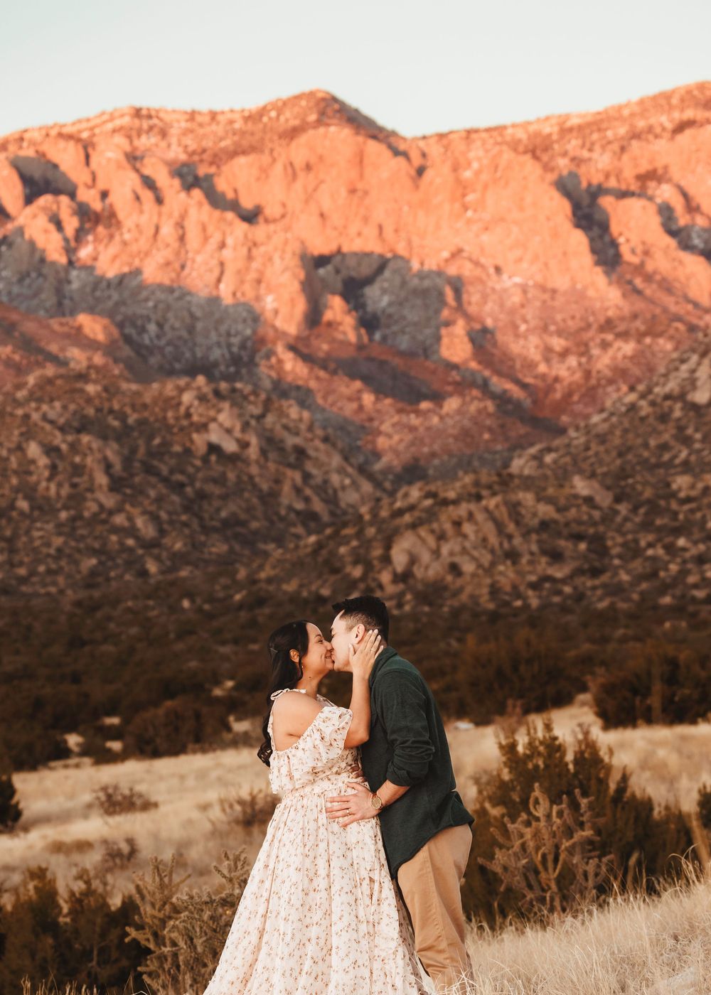 Couple sharing a romantic kiss in a golden field with stunning red-lit mountains during a maternity photoshoot.