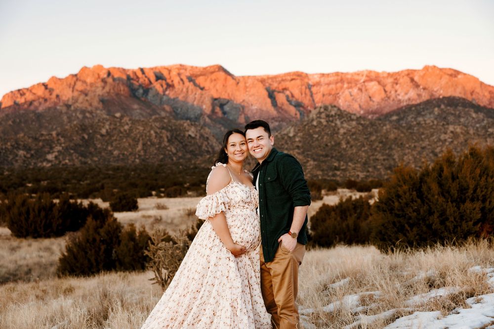 Smiling couple posing together in a field at sunset with red Sandia Mountains in the background during maternity shoot.