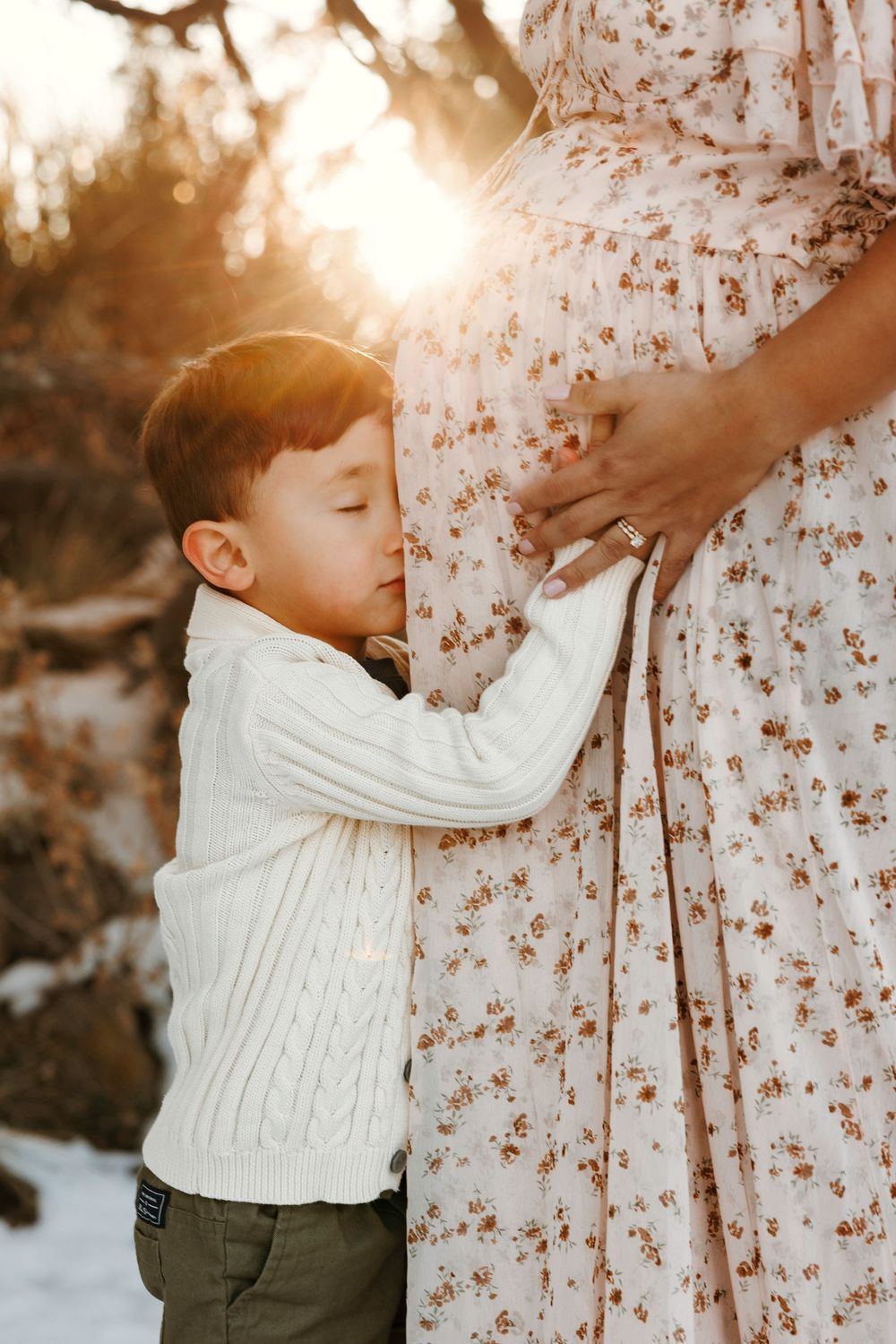 A young boy kisses his pregnant mothers belly in a golden sunset outdoor setting.