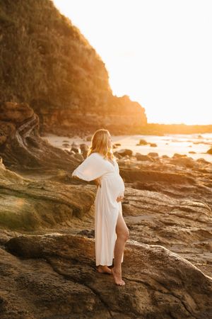 Woman in flowing white dress stands on coastal rocks at sunset while golden light illuminates the rocky shoreline.