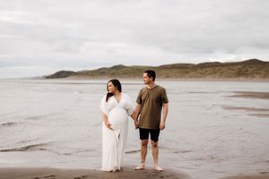 A couple stands together on a sandy beach with rolling hills in the background under an overcast sky.