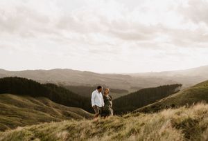 Couple stands together on hilltop overlooking mountain valley at sunset.