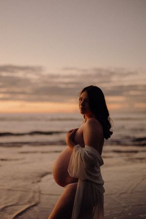 Silhouette of pregnant figure in flowy dress standing on beach at sunset with waves and cloudy sky in background.