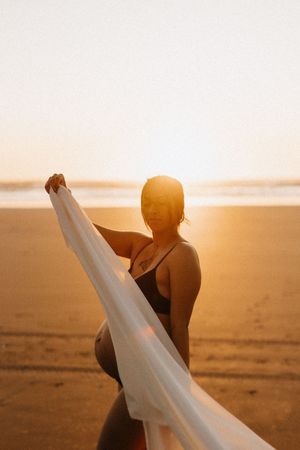 Woman in black swimwear holding white fabric on beach during golden hour.