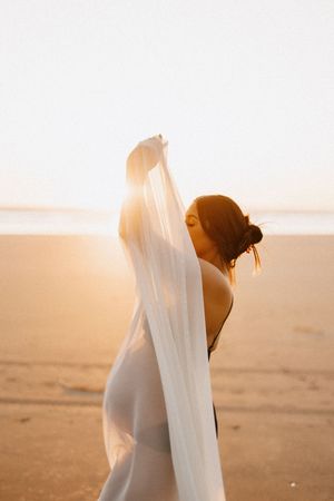 Pregnant woman in white dress holding sheer fabric while standing on sandy beach at sunset.
