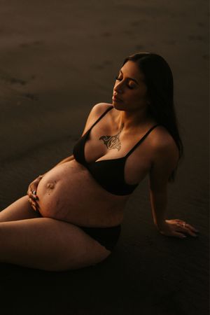 Maternity beach photo of woman in black swimwear sitting on dark sand at dusk.