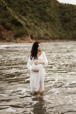 A pregnant woman in a white dress stands in shallow ocean waters near a rocky coastline.