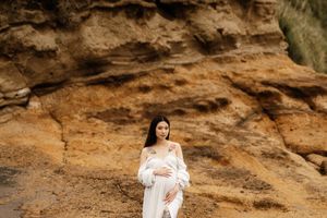 An expectant mother in a white dress poses against a dramatic rocky cliff backdrop.