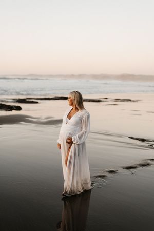 Expectant mother in flowing white dress stands reflectively on wet sand during golden hour.