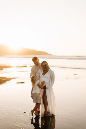 A couple embraces on a serene beach at sunset with their feet in the shallow water's reflection.
