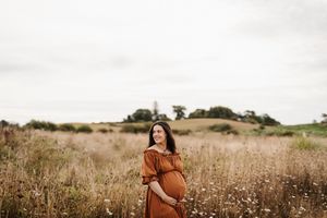 Expectant mother in rust-colored dress standing in a golden field at sunset.