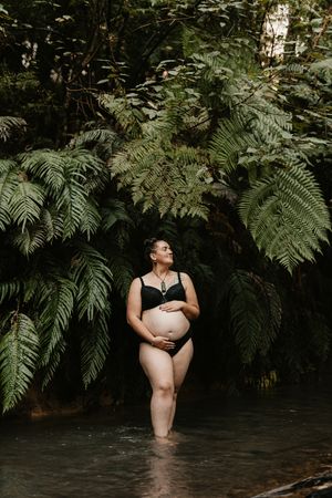 Maternity photo of woman in black swimsuit surrounded by large silver ferns near stream.