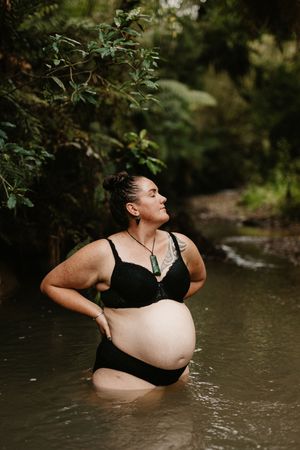 Pregnant woman in black swimwear stands in forest stream during outdoor photoshoot.