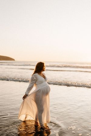 Ethereal maternity photo of an expectant mother in a flowing white dress standing at the ocean's edge during sunset.