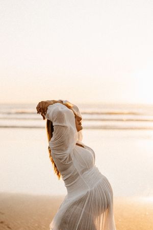 Artistic silhouette of pregnant woman in white dress posing on beach at golden hour.