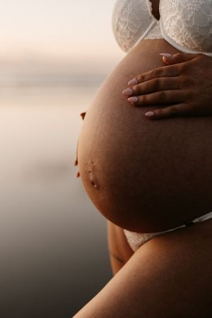 Close-up shot of pregnant belly with hands gently resting on it against a sunset beach backdrop.