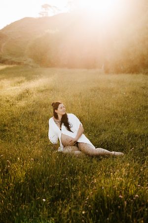 Woman in white dress sits peacefully in sunlit meadow with mountains in background.