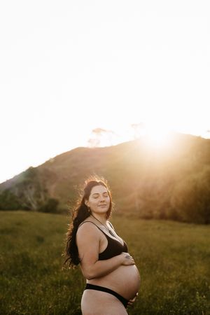Pregnant woman in black swimwear poses against golden sunset in grassy hillside.