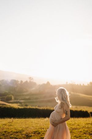 Person in a white dress stands in a sunlit meadow during golden hour with rolling hills in the background.